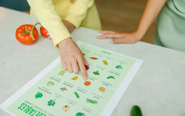 Close-up of hands pointing to a vegetable nutrition chart with fresh tomatoes on the table.