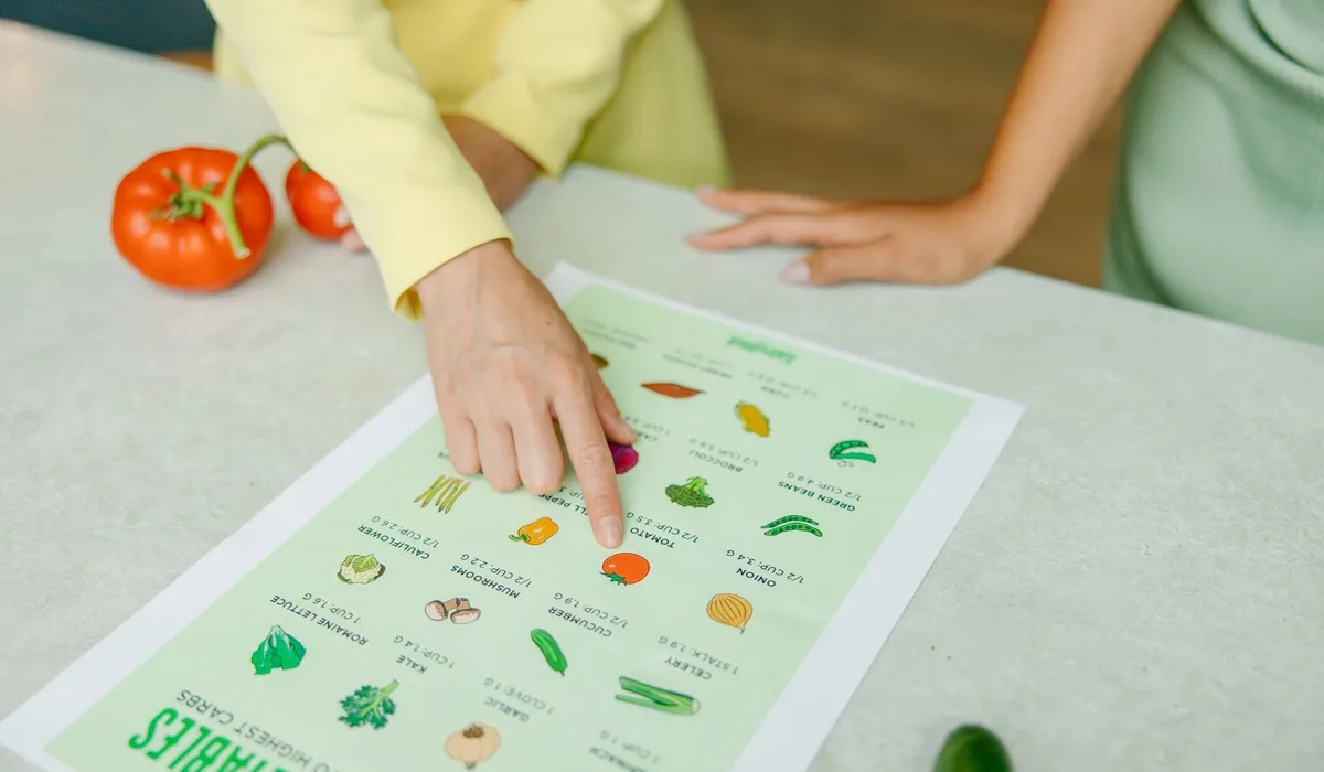 Close-up of hands pointing to a vegetable nutrition chart with fresh tomatoes on the table.